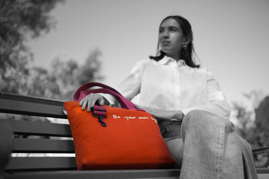 Kaahu canvas tote bag with bold red fabric and inspirational quote "Be your own light", carried by woman in white shirt and jeans on park bench, lifestyle outdoor scene.